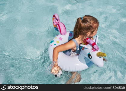 Little girl playing in a swimming pool with inflatable rubber ring toy in the shape of unicorn. View from above of kid in a turquoise water swimming in rubber colorful ring. Little girl playing in a swimming pool with inflatable ring toy in the shape of unicorn