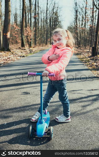 Little girl playing in the park, riding a scooter, having fun on sunny autumn day. Real people, authentic situations