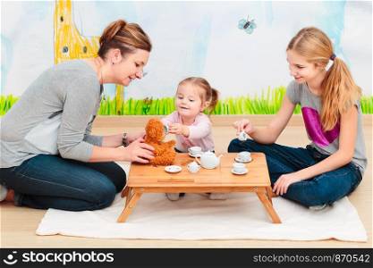 Little girl playing with her mother and elder sister at tea party using childs tea set