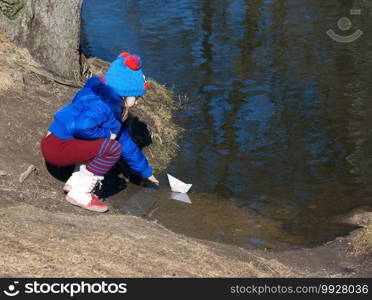 little girl playing with paper boat on the river on sunny spring day
