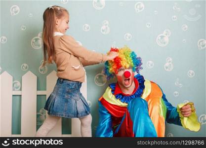 Little girl pulls clown hair. White fence decoration on the background.