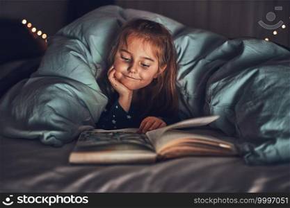 Little girl reading book and watching pictures in bed before going to sleep. Reading stories before sleep. Bedtime stories for child