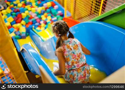Little girl riding on plastic kids slide, playground in entertainment center. Play area indoors, playroom