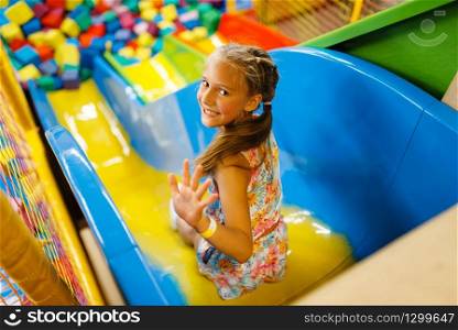 Little girl riding on plastic kids slide, playground in entertainment center. Play area indoors, playroom. Little girl riding on plastic kids slide, playroom