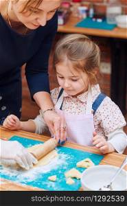 Little girl rolling the dough with her mom&rsquo;s help for the cookies. Cutting the dough to heart shapes. Kid taking part in baking workshop. Baking classes for children, aspiring little chefs. Learning to cook. Combining and stirring prepared ingredients. Real people, authentic situations