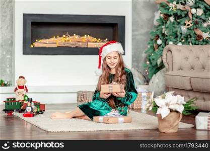 Little girl Santa Claus hat with gifts under Christmas tree sitting by the fireplace, unpacks gifts. Merry Christmas.. Little girl Santa Claus hat with gifts under Christmas tree sitting by the fireplace, unpacks gifts.