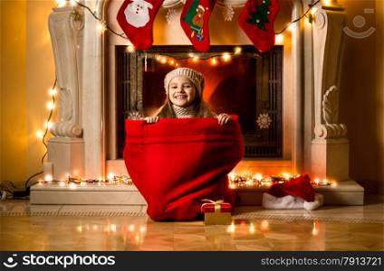 Little girl sitting in big red sack at room decorated for Christmas