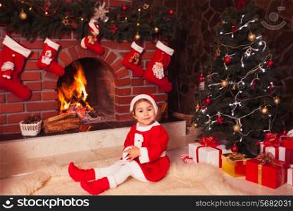 Little girl sitting near fireplace and christmas tree with gift boxes
