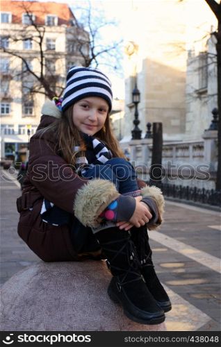 little girl sitting on a stone sphere at clear autumn morning. City landscape at the background
