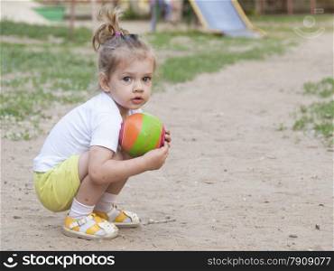 Little girl sitting on his haunches with a ball on the Playground. Summer evening time