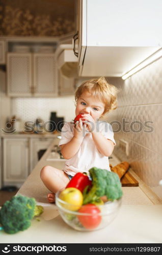 Little girl sitting on the table and eats tomato. Female baby tasting vegetables from the bowl on the kitchen. Child tastes vegetarian food