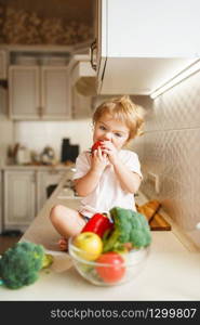 Little girl sitting on the table and eats tomato. Female baby tasting vegetables from the bowl on the kitchen. Child tastes vegetarian food