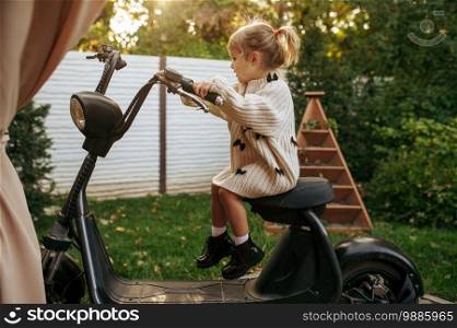 Little girl sitting on vintage scooter in the garden. Female child poses on motor vehicle on backyard. Kid having fun on playground outdoors, happy childhood. Little girl sitting on vintage scooter in garden