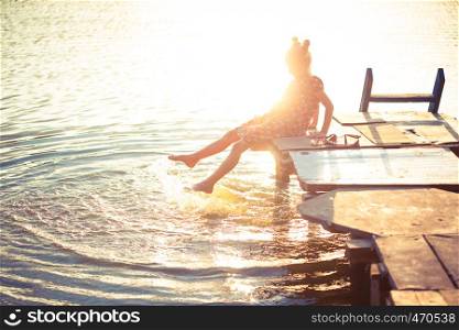 little girl splashing at the lake sitting on a wooden pier at the sunset time