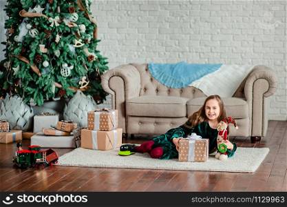 Little girl under the Christmas tree in Santa Claus hat with gifts under the Christmas tree by the fireplace. Unpacking gifts. Merry Christmas.. Little girl under the Christmas tree in Santa Claus hat with gifts under the Christmas tree by the fireplace.