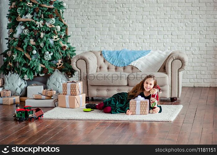 Little girl under the Christmas tree in Santa Claus hat with gifts under the Christmas tree by the fireplace. Unpacking gifts. Merry Christmas.. Little girl under the Christmas tree in Santa Claus hat with gifts under the Christmas tree by the fireplace.