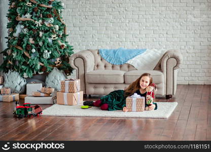 Little girl under the Christmas tree in Santa Claus hat with gifts under the Christmas tree by the fireplace. Unpacking gifts. Merry Christmas.. Little girl under the Christmas tree in Santa Claus hat with gifts under the Christmas tree by the fireplace.