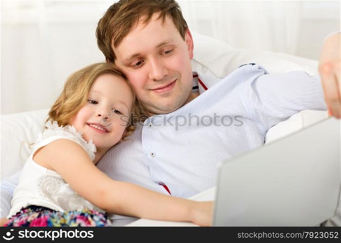 Little girl using laptop sitting on the sofa with father and leaning on his shoulder. Happy dad and daughter with laptop