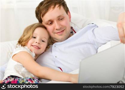 Little girl using laptop sitting on the sofa with father and leaning on his shoulder. Happy dad and daughter with laptop