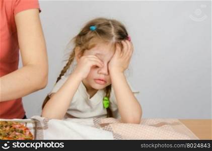 Little girl wearily rubbing his eyes at the kitchen table while Mom cooks