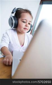 Little girl wearing headphones while looking at laptop at table in house