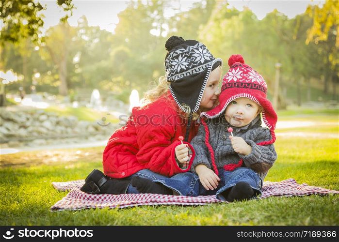 Little Girl Whispers A Secret to Her Baby Brother Wearing Winter Coats and Hats Sitting Outdoors at the Park.