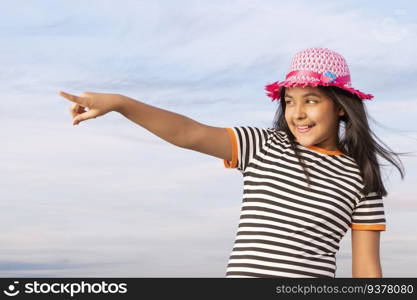 Little girl with hat standing against blue sky and pointing away