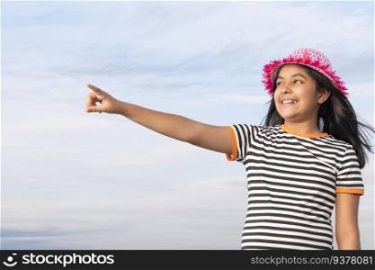 Little girl with hat standing against blue sky and pointing away