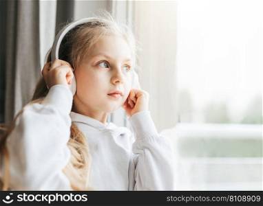 Little girl with headphones indoors at home, sitting on window sill and listening music