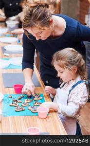 Little girl with her mom&rsquo;s help decorating baked cookies with colorful sprinkle and icing sugar. Kid taking part in baking workshop. Baking classes for children, aspiring little chefs. Learning to cook. Combining and stirring prepared ingredients. Real people, authentic situations