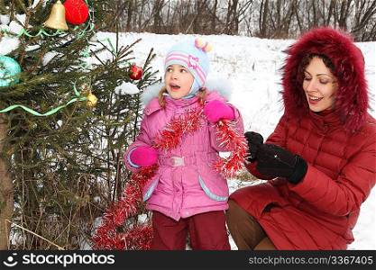 little girl with her mother wearing winter jacket is decorating christmass tree.