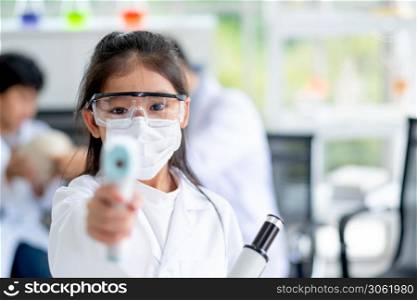 Little girl with hygiene mask and protective eye glasses show infrared thermometer and look like she use for measure temperature of someone in laboratory or class room.