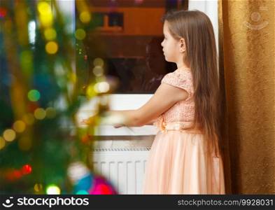little girl with long brown hair and in pink dress standing at the window at Christmas