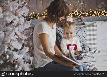 Little girl with mother reading a book sitting under the Christmas tree.. Mother year-old daughter read the book in Christmas decorations 7110.