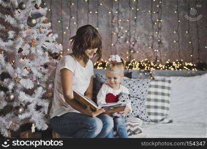 Little girl with mother reading a book sitting under the Christmas tree.. Mother year-old daughter read the book in Christmas decorations 7107.