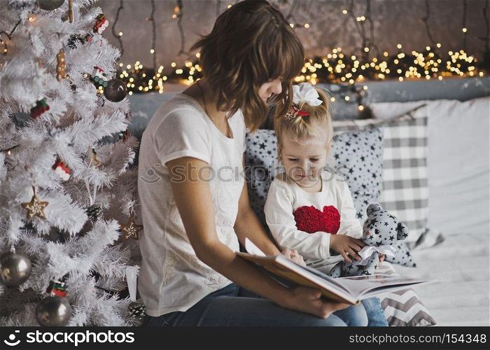Little girl with mother reading a book sitting under the Christmas tree.. Mother year-old daughter read the book in Christmas decorations 7109.
