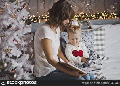 Little girl with mother reading a book sitting under the Christmas tree.. Mother year-old daughter read the book in Christmas decorations 7109.