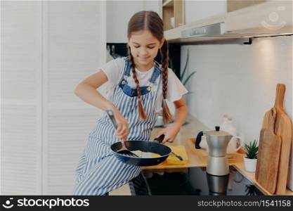 Little girl with pigtails wears apron, learns to cook, poses near stove, prepares fried eggs for breakfast, helps parents with cooking, busy at modern kitchen. Children, culinary, food concept