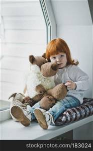 Little girl with red hair plays with a toy.. Girl with a toy sitting on the windowsill 4392.