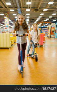 Little girlfriends rides on scooters in the entertainment center. Female children leisures on holidays, childhood happiness, happy kids on playground. Little girlfriends rides on scooters, playground