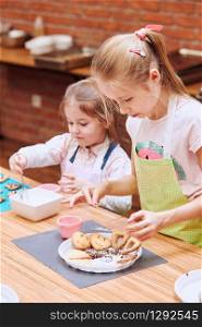 Little girls decorating baked cookies with colorful sprinkle and icing sugar. Kids taking part in baking workshop. Baking classes for children, aspiring little chefs. Learning to cook. Combining and stirring prepared ingredients. Real people, authentic situations