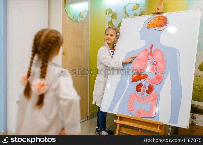 Little girls in uniform playing doctor at the poster with human organs, playroom. Kids plays medicine worker in imaginary hospital, profession learning, childish dream. Girls playing doctor at poster with human organs