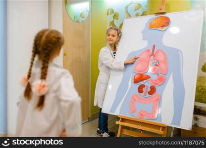 Little girls in uniform playing doctor at the poster with human organs, playroom. Kids plays medicine worker in imaginary hospital, profession learning, childish dream. Girls playing doctor at poster with human organs