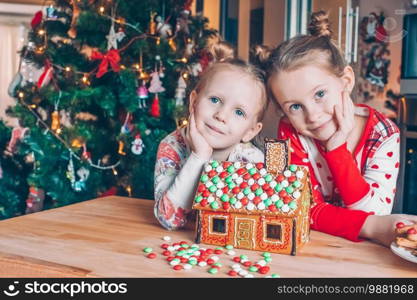 Little girls making Christmas gingerbread house at fireplace in decorated living room. Kids playing with ginger bread under Christmas tree. Baking and cooking with children for Xmas at home. - Image. Little girls making Christmas gingerbread house at fireplace in decorated living room.