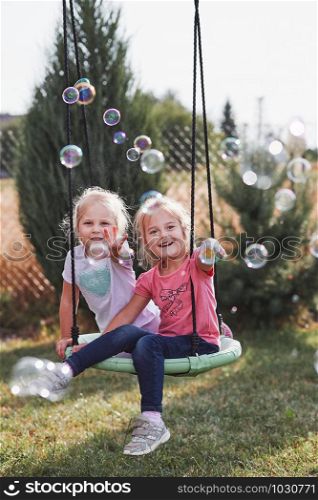 Little girls playing with soap bubbles outdoors sitting on swing on summer day. Real people, authentic situations