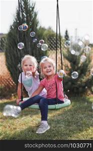 Little girls playing with soap bubbles outdoors sitting on swing on summer day. Real people, authentic situations