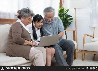 Little granddaughter teach senior elder to surf internet using computer and technology& modern lifestyle.Happy asian grandparent with little young cute grandchild sitting on sofa playing laptop together.