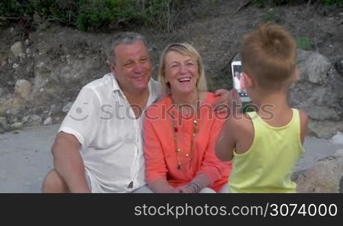 Little grandson making photo of happy grandmother and grandfather outdoor. He using smart phone to take a great shot