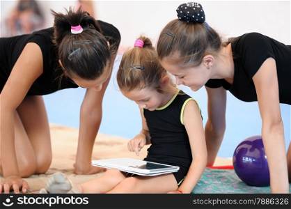 Little gymnast girl using tablet PC sitting on the floor while other two looking on it