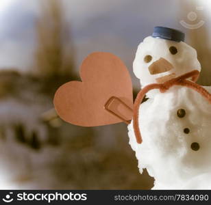 Little happy christmas snowman in blue screw top as hat red scarf and with clip red heart love symbol toothpick outdoor. Winter season seasonal specific valentine&#39;s day. Blue sky background. Sepia tone.